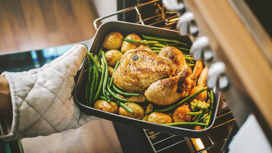a pan with a whole roasted chicken and green beans being pulled from an oven