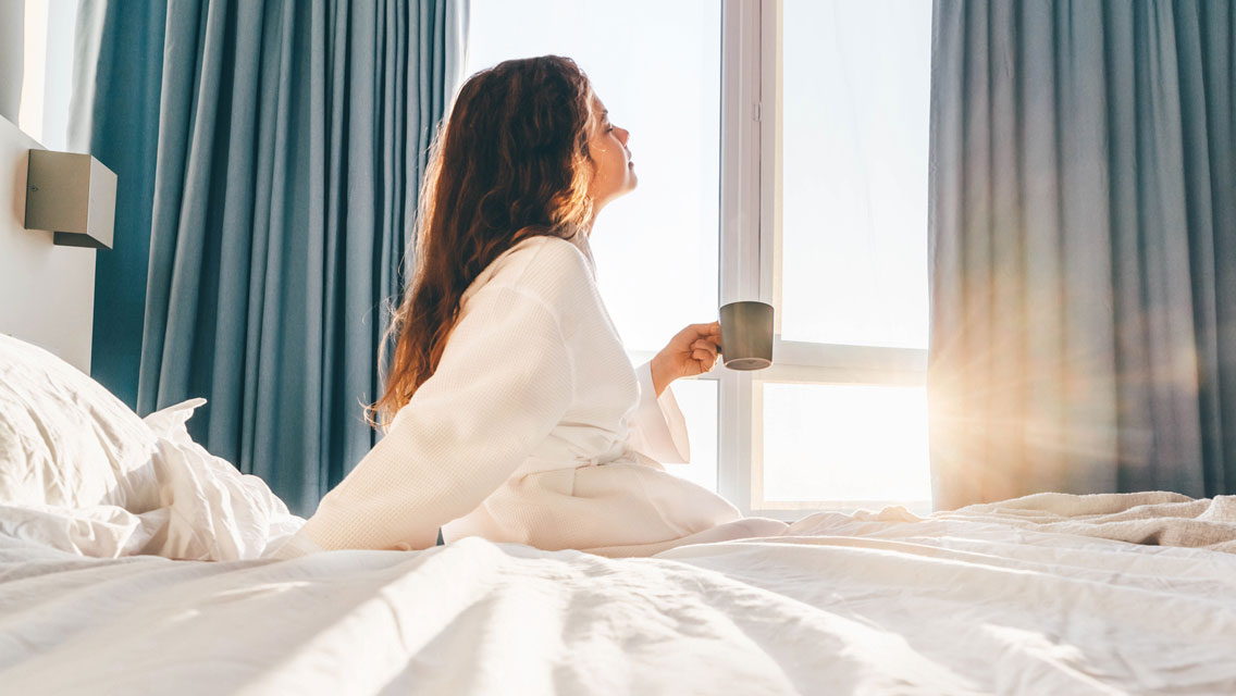 a woman looks out a window from her bed with a cup of coffee