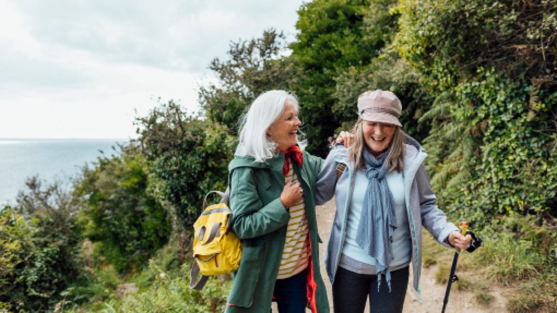 two senior women hike and laugh on a mountain side trail