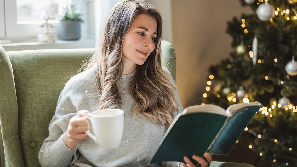 a woman holds a coffee mug while reading by a Christmas tree