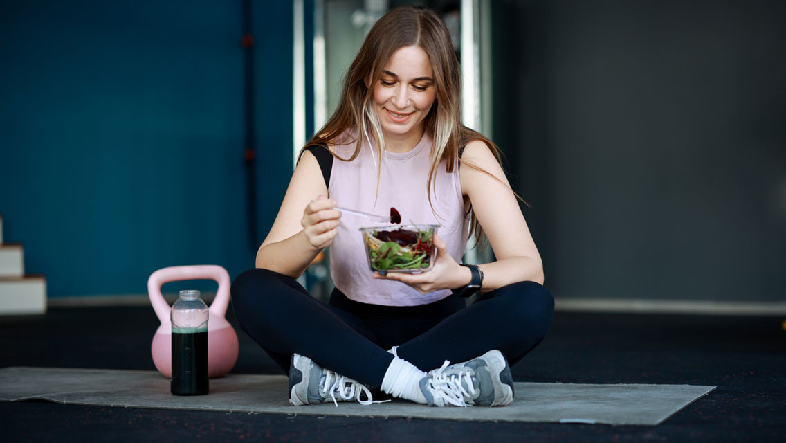a woman eats a salad on her yoga mat with a kettlebell and water bottle next to her