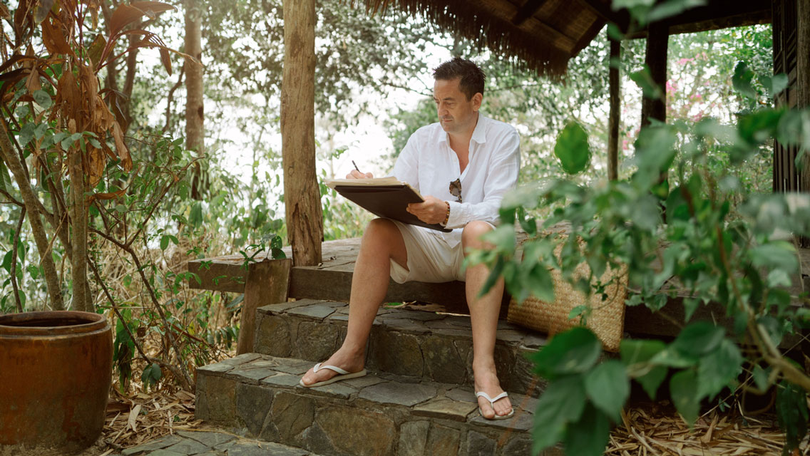 a man writes while sitting on the front step of a retreat center in a tropical area