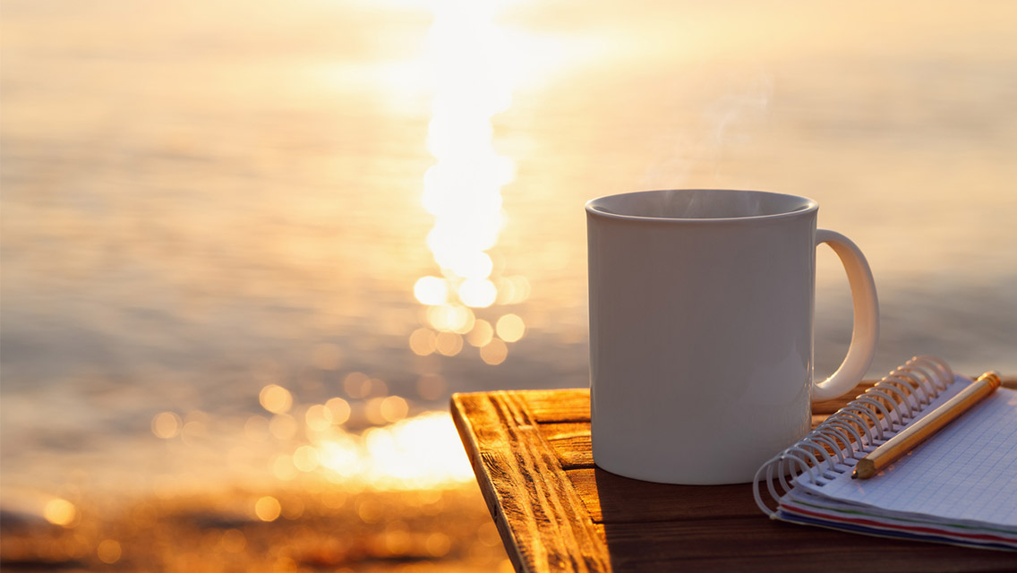 a steaming cup of coffee sits next to a journal at sunrise