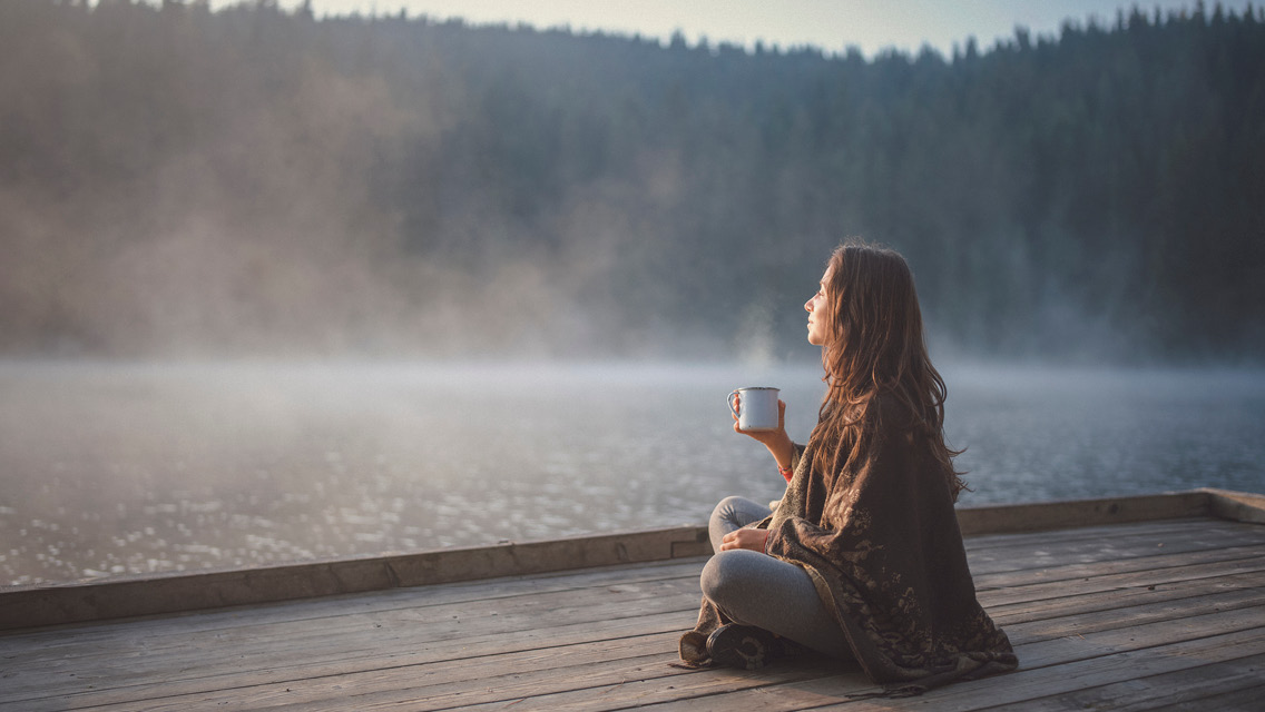 a woman sits on a dock with a steaming cup of coffee