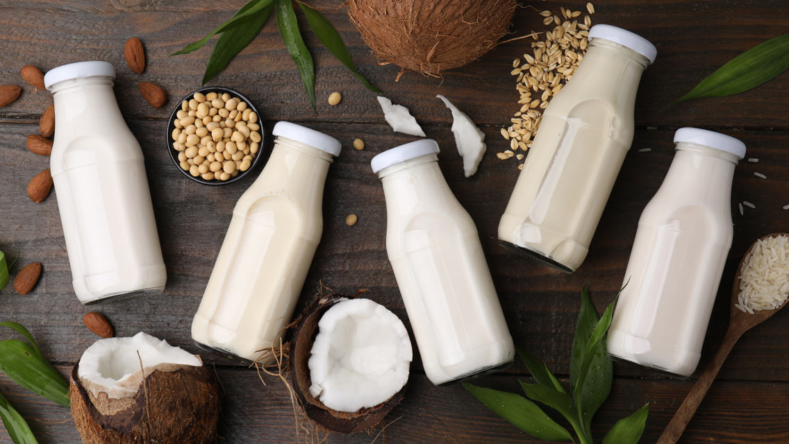 Different types of vegan milk in bottles with ingredients and green leaves on wooden table