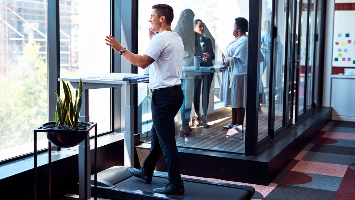 a young business professional talks on his phone while walking on a desk treadmill