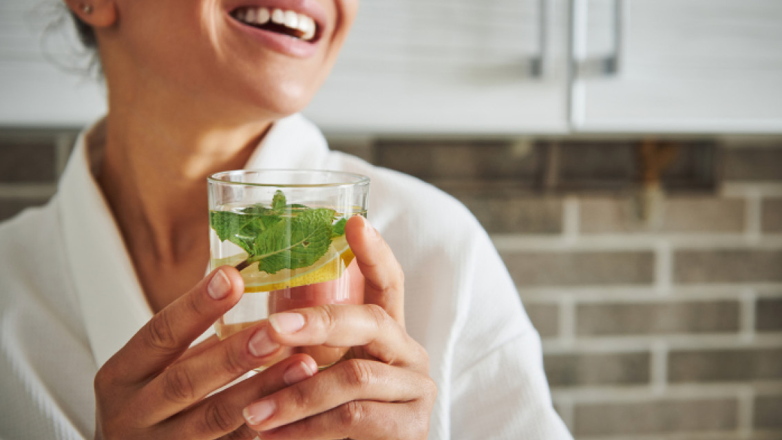 a woman drinks a glass of water with a sprig of mint in it