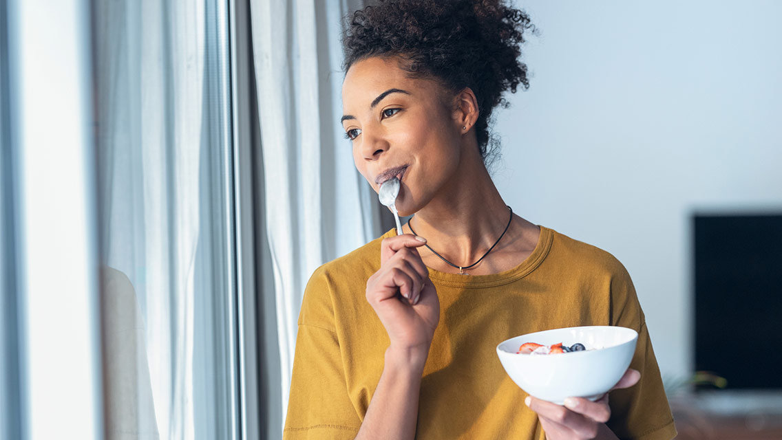 Woman eating bowl of yogurt and fruit next to window