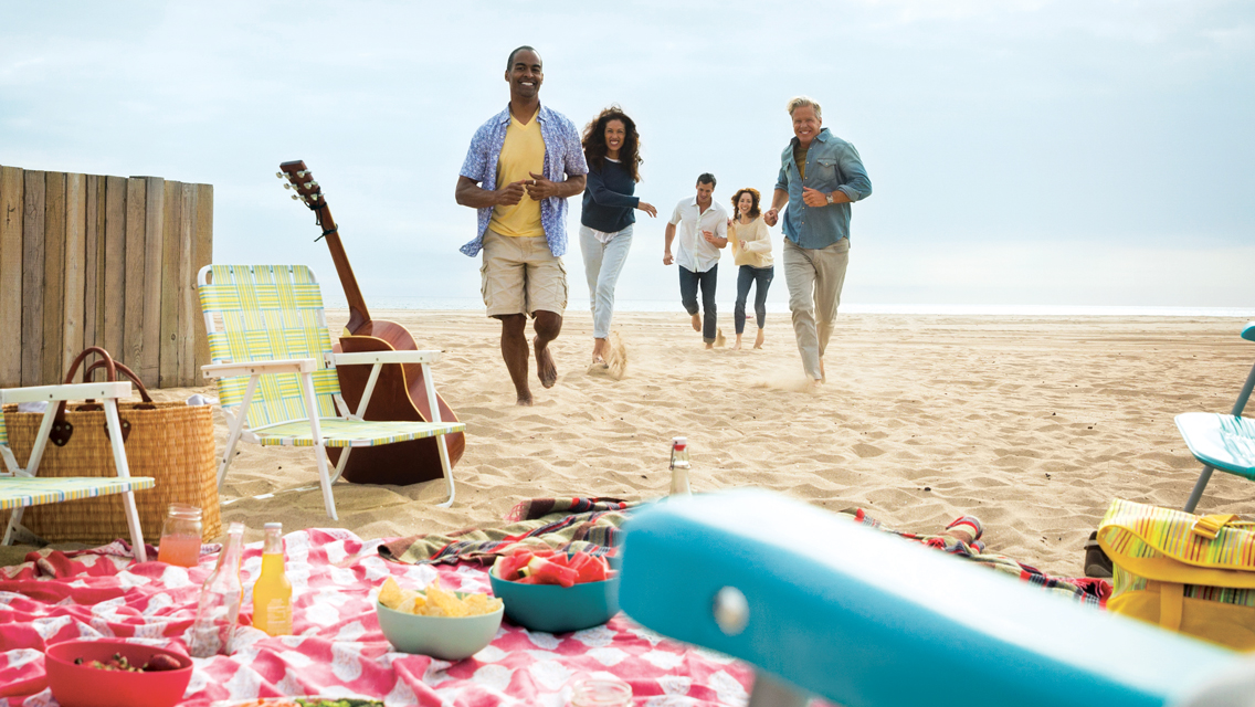 a group of friends run towards their picnic set up on a beach