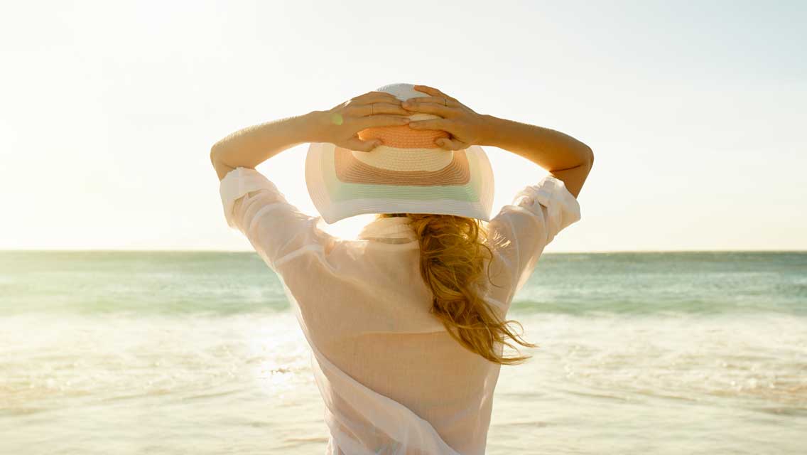 a woman stands holding her hat on on a sunny beach