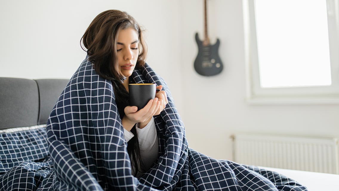 A woman wrapped in a blanket bends over a cup of tea.