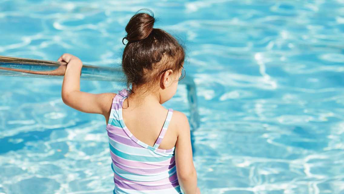 A young girl holding onto a rail while walking into the shallow end of a pool.
