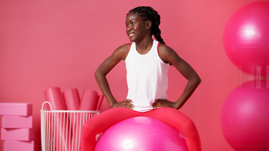 A young girl in a room with a lot of pink play items, including an exercise ball and swimming noodles.