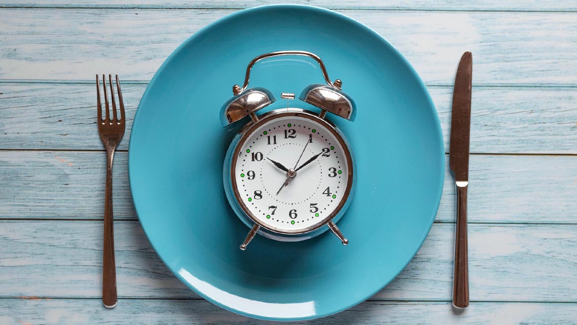Intermittent fasting concept with clock on plate, fork and knife on wooden table