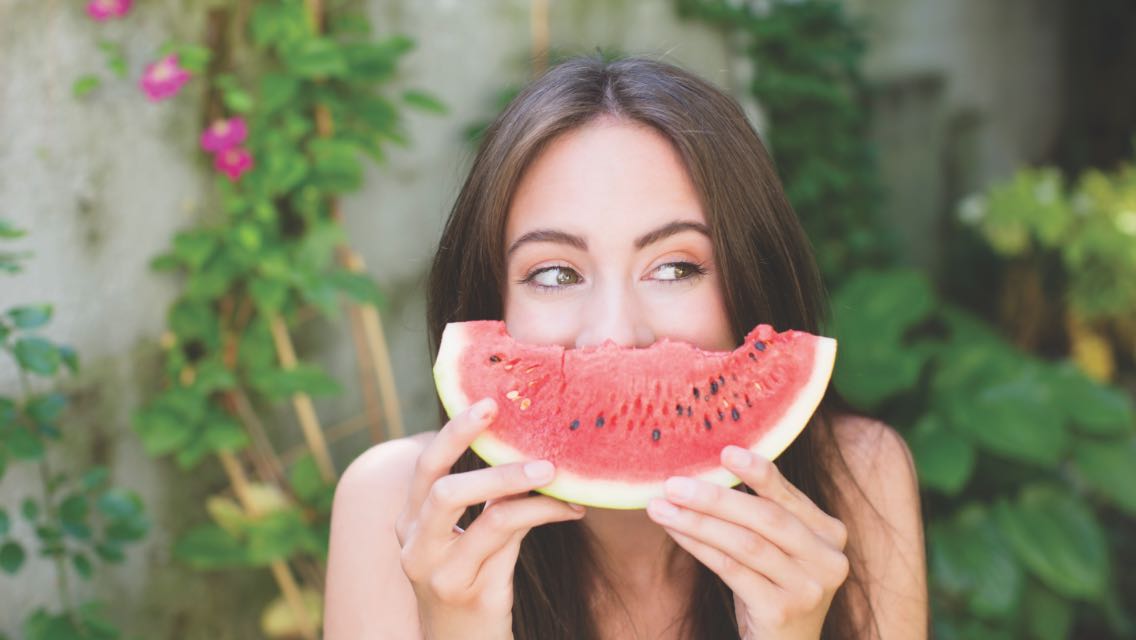 Woman with piece of watermelon covering her mouth like a smile.
