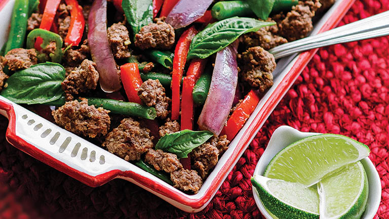A casserole dish of Thai Basil beef with red onions, red peppers, and lime wedges