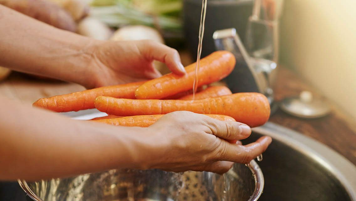 A close up of a person washing carrots over a colliander in the sink.