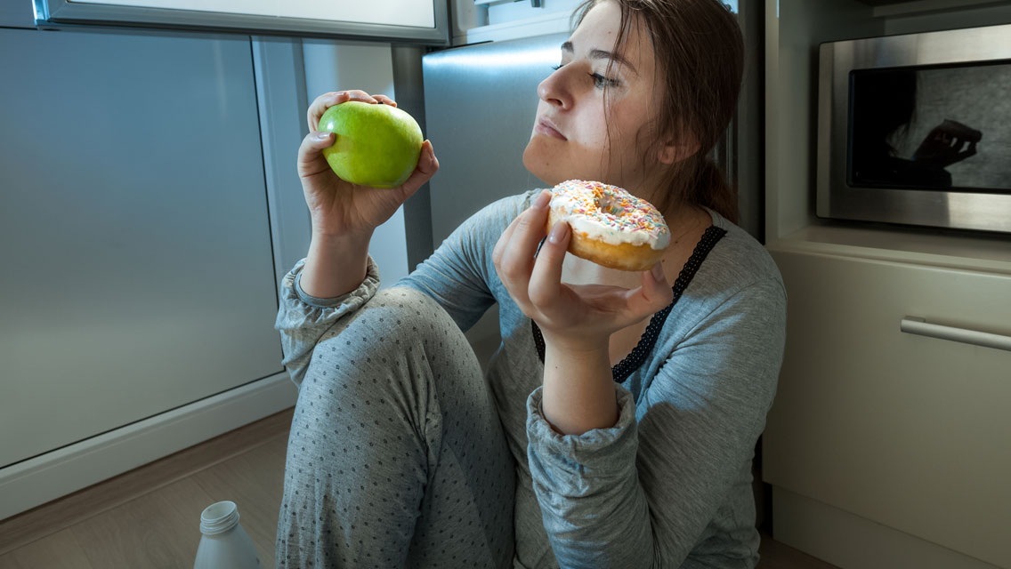 a woman sits by her refrigerator at night with an apple in one hand and donut in the other