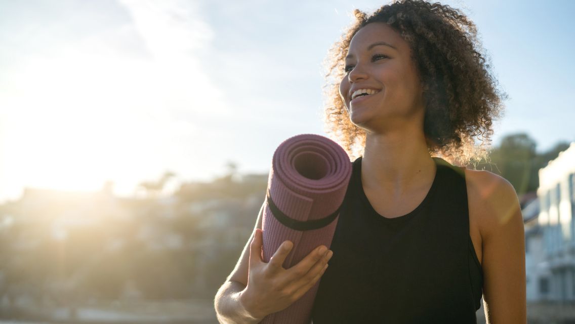 A woman smiling while walking outside holding a yoga mat.