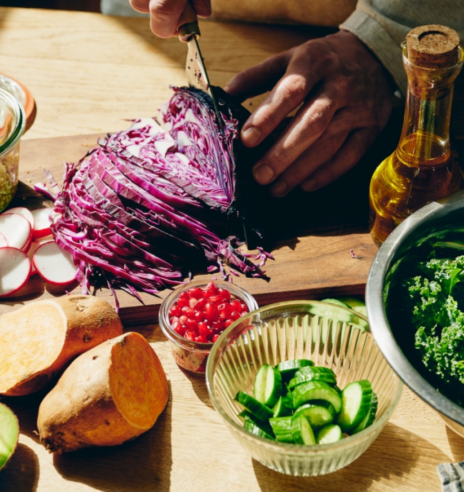 A close up of a hand chopping red cabbage on a table alongside other foods including kale, cucumber, sweet potatoes, and radishes.