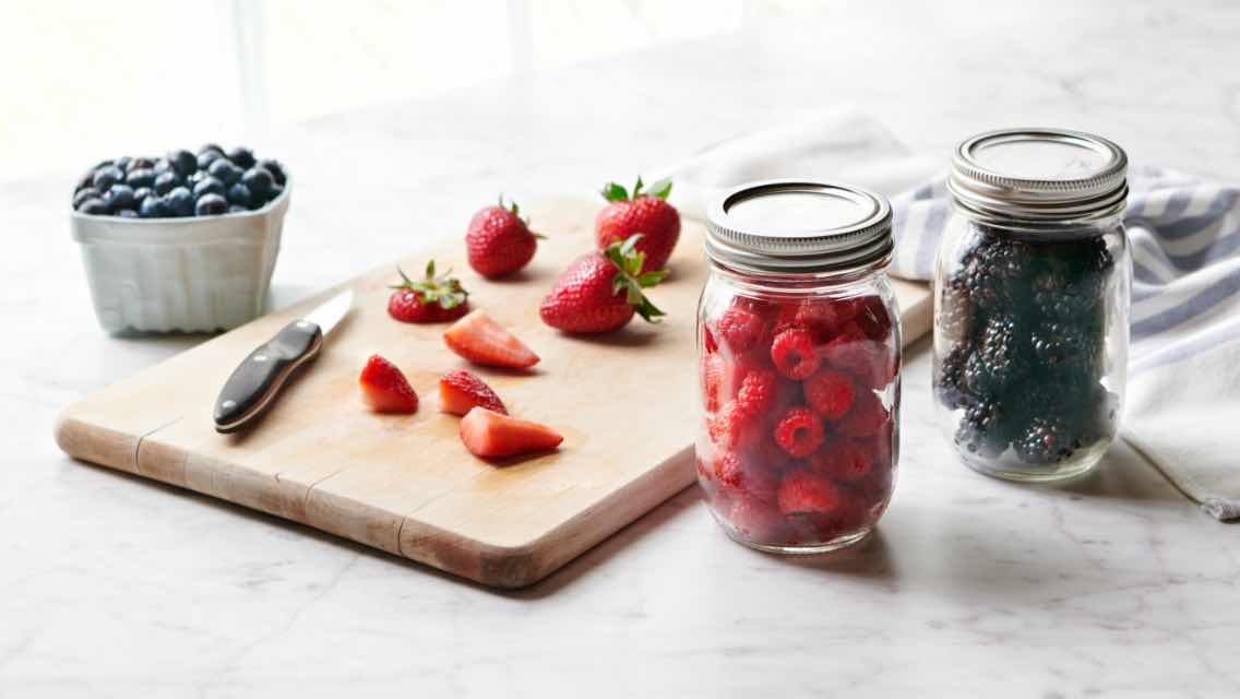 Two mason jars, one filled with raspberries, one with blueberries, a cutting board with sliced strawberries, and a bowl of blueberries.