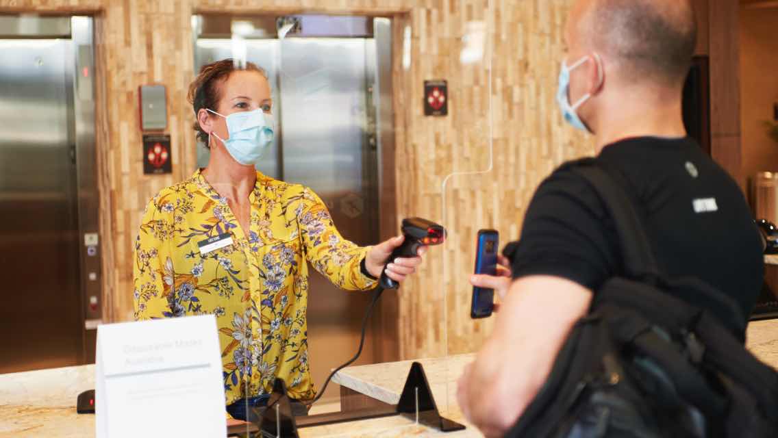 A woman with a face mask on checking someone in through a plexiglass window at a health club.