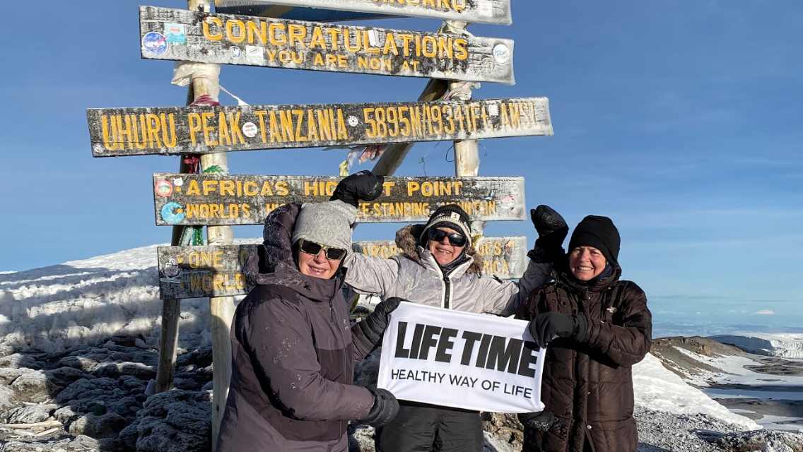 Three women on the top of Mount Kilimanjaro holding a sign that says "Life Time."