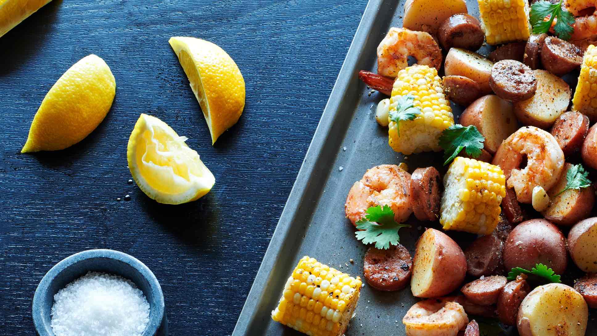 Corn, sausage, shrimp, and potatoes on a sheet pan, with a bowl of sea salt and a few lemon wedges on the table next to it.