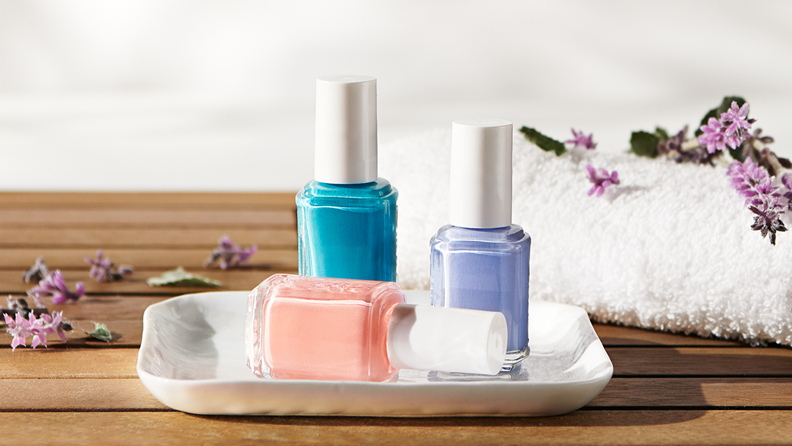 Bottles of coral, purple, and blue nail polish on a dish on a surface next to a towel.