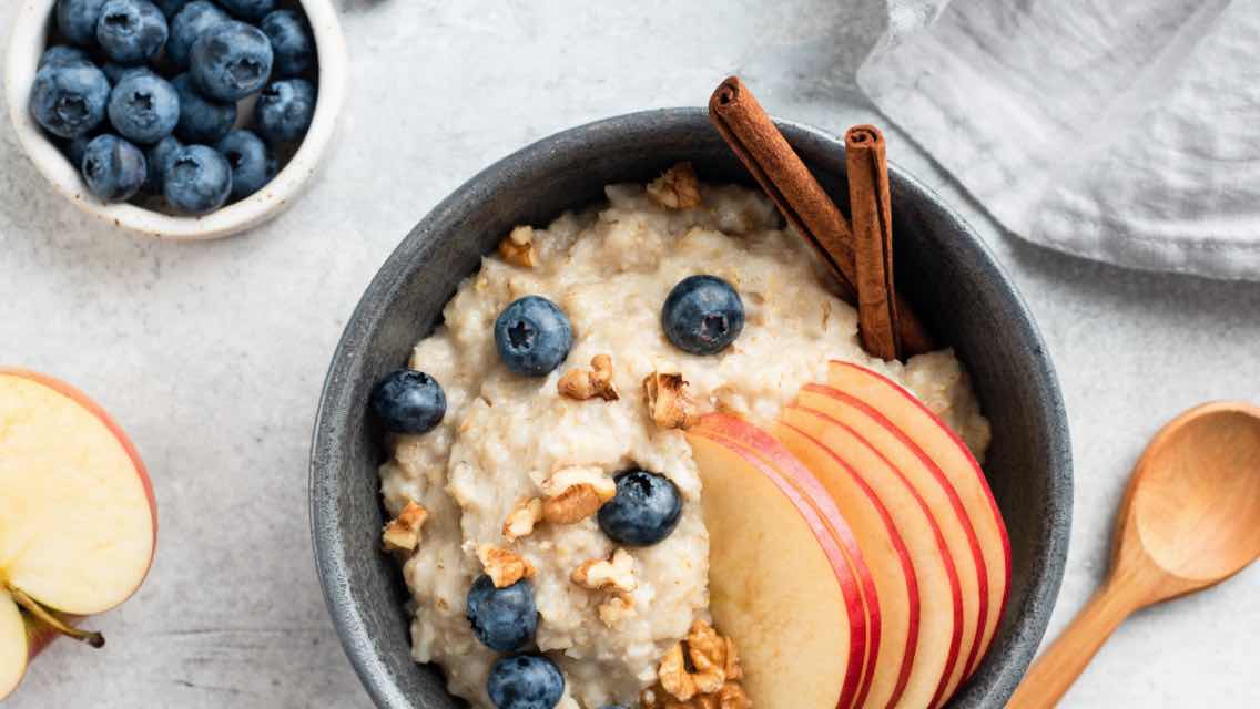 A bowl of oatmeal topped with apple slices, blueberries, walnuts, and cinnamon sticks.