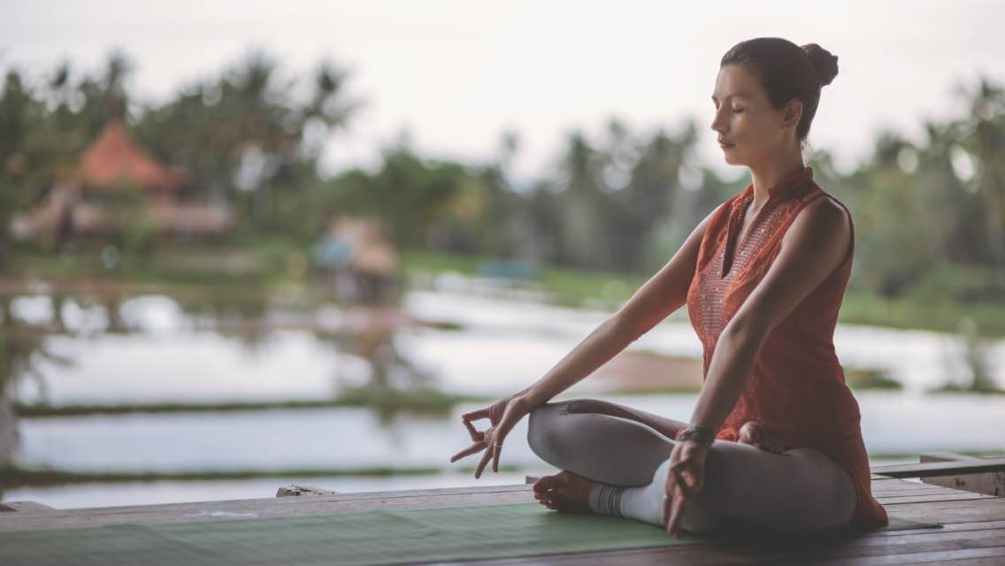 A woman outside in a serene setting sitting cross-legged in a meditative pose.