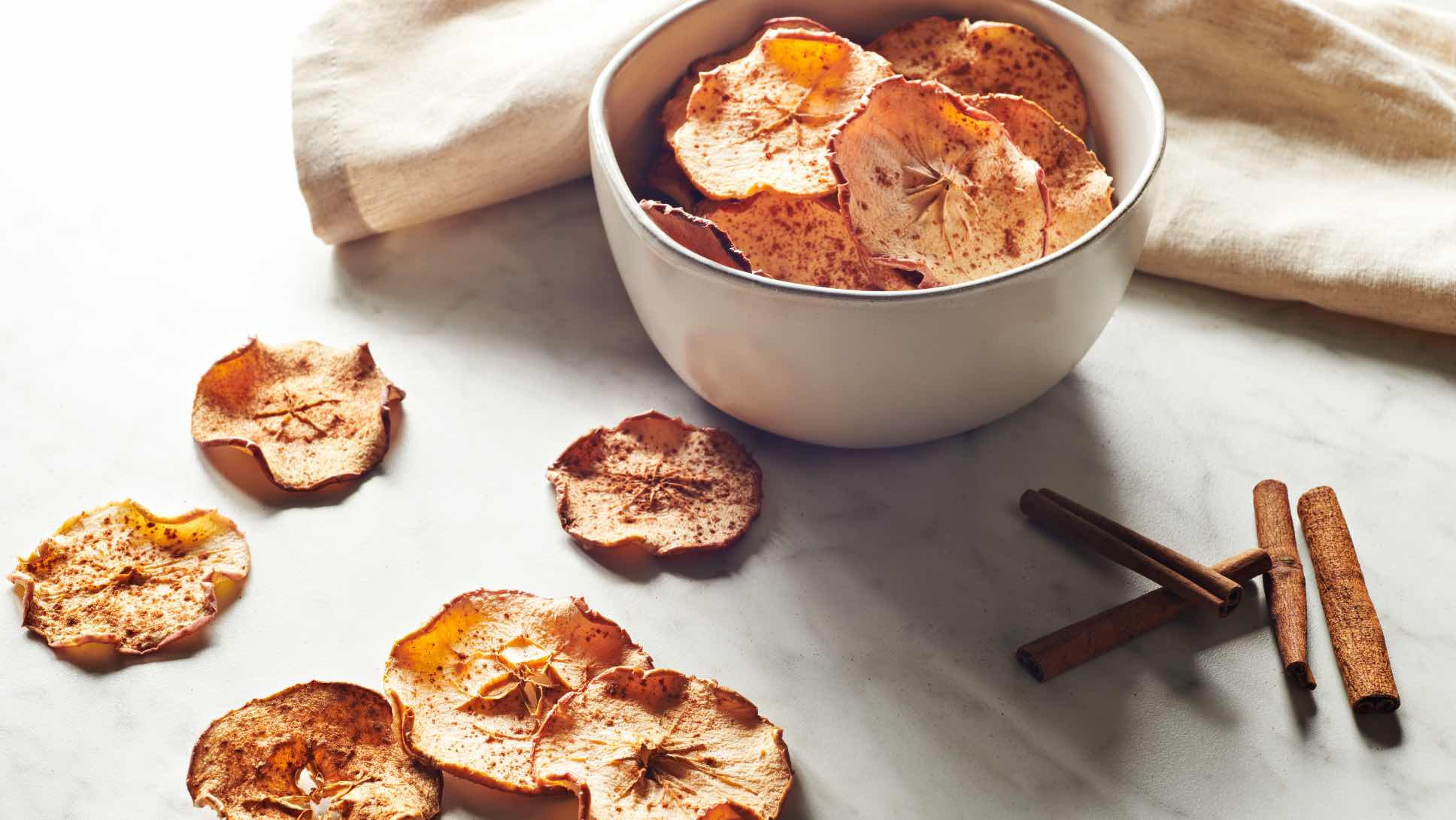 A bowl of cinnamon apple chips with a few apple chips and cinnamon sticks on a counter next to the bowl.
