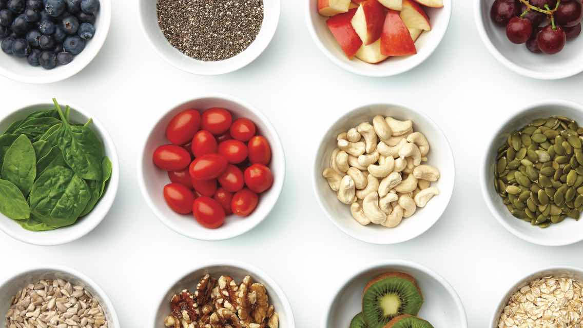 A variety of food in bowls, including spinach, tomatoes, nuts and seeds, apples, kiwi, and blueberries.
