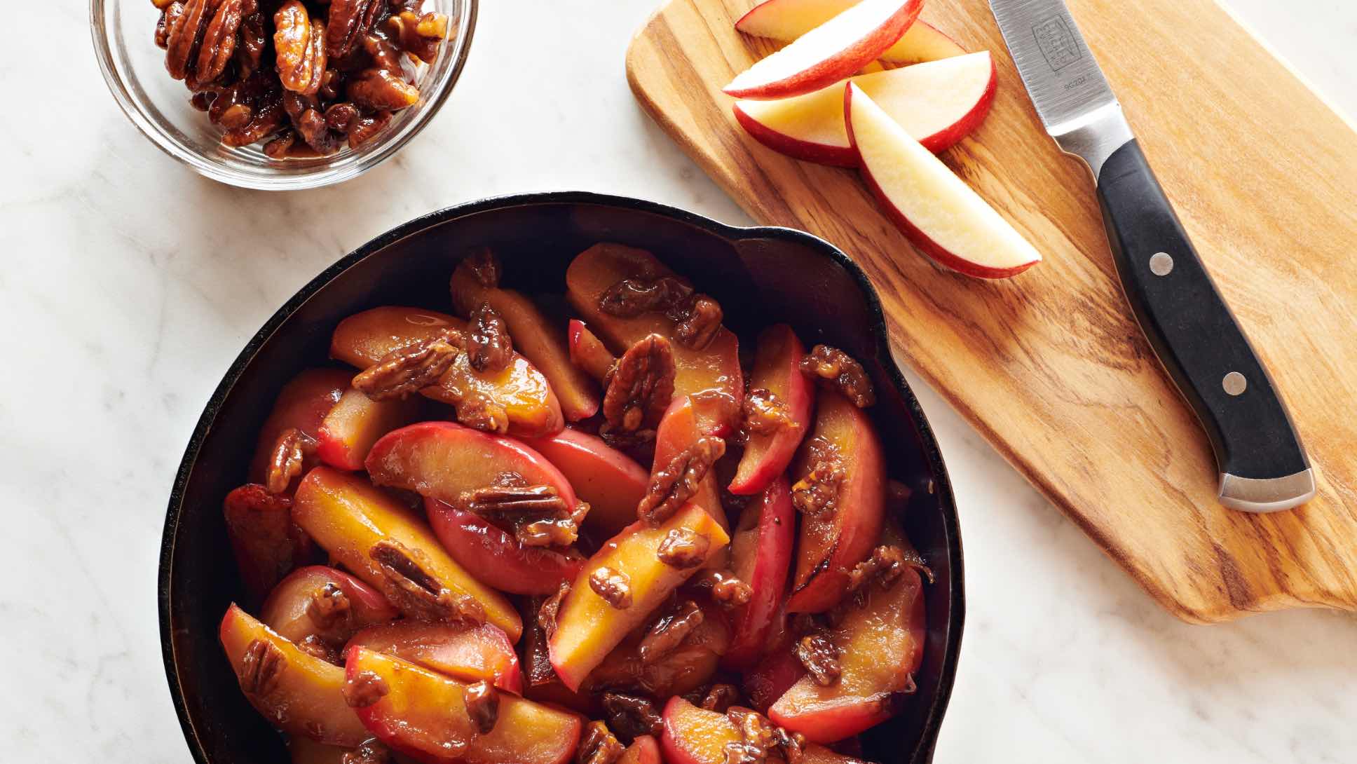 A cast-iron skillet of sauteed apples with maple-glazed pecans next to slices of apple and a bowl of pecans.