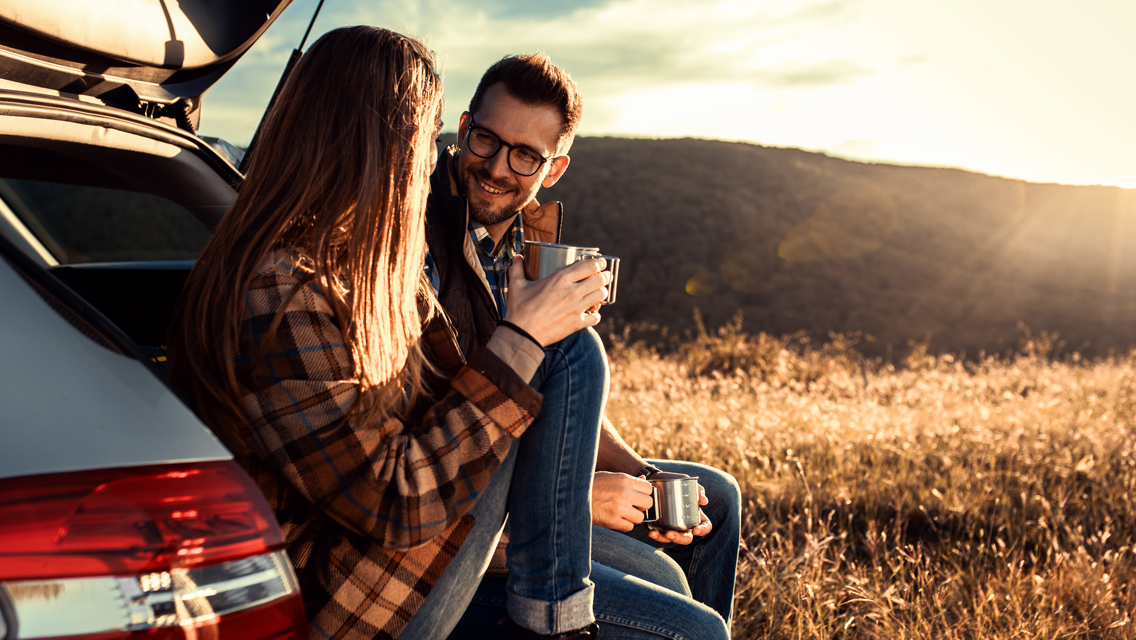 a couple talking while sitting in the trunk of their suv drinking coffee