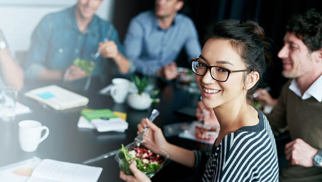 Smiling co-workers, one eating a salad, are shown.