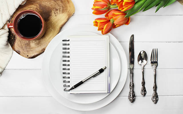Overhead shot of a journal over empty plates with a cup of coffee and a bouquet of springtime tulip flowers over white wood table top.