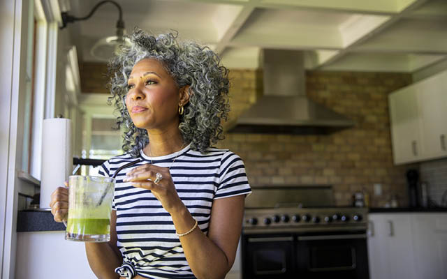 A woman drinks a green smoothie while she smiles out the window.