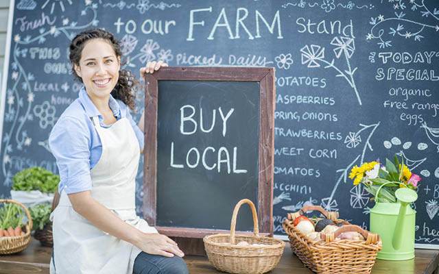 A woman at a farmer's market holds up a "BUY LOCAL" sign.
