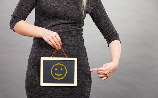 A woman holds a smiley sign over her pelvic region.