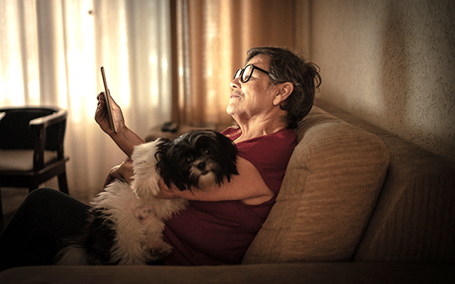 A woman sitting on a couch holds a dog and a phone.