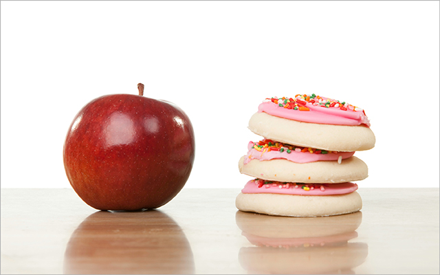 An apple sits next to a stack of pink frosted cookies.
