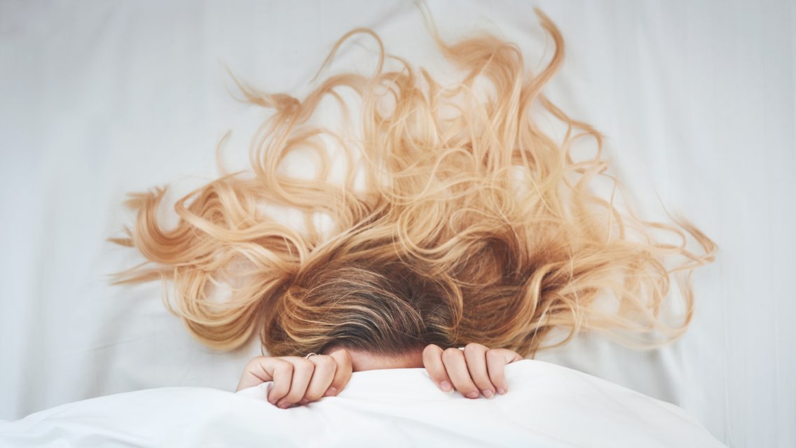 the top of a blond woman's head peers out from under a bed sheet