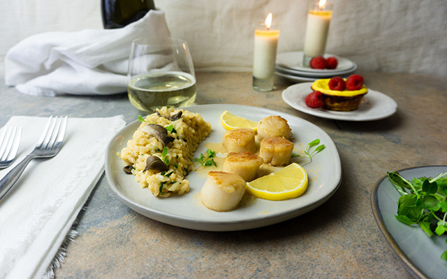 An elegant meal of scallops, risotto, and custard tart is presented on a table.