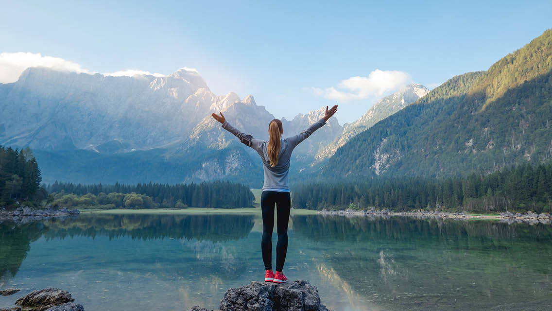a woman stands looking out on a mountain lake with her arms outstretched