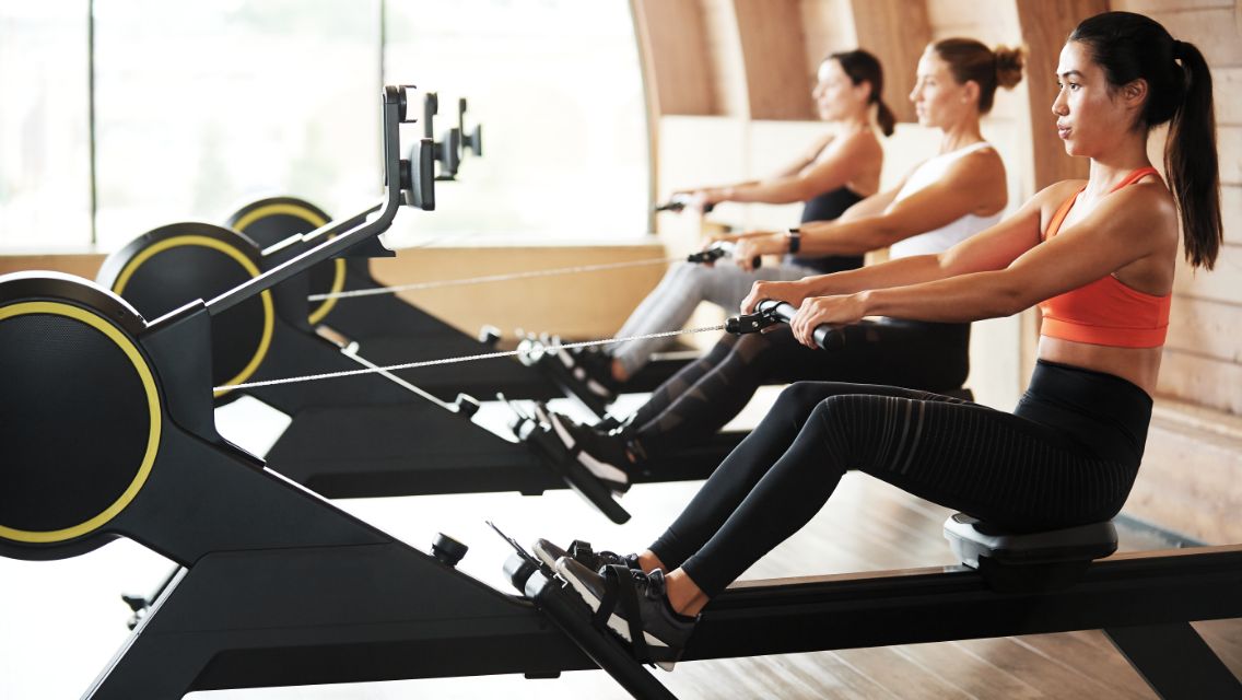 Three women on rower machines inside of a Life Time health club.