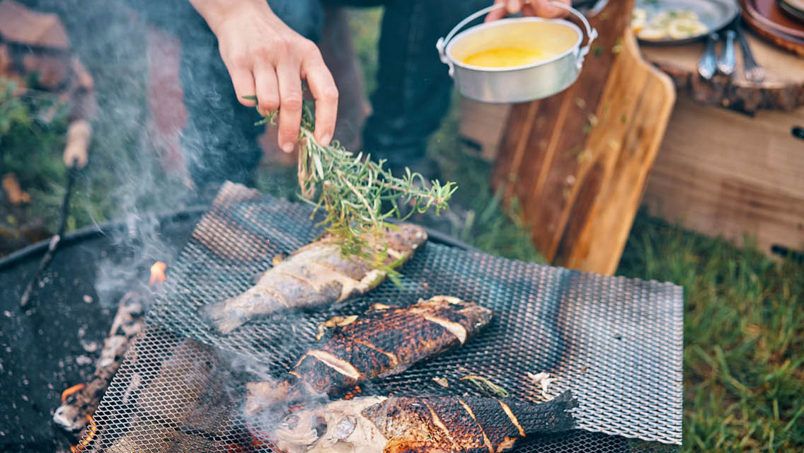 a person uses sprigs of rosemary to brush butter over fish being grilled