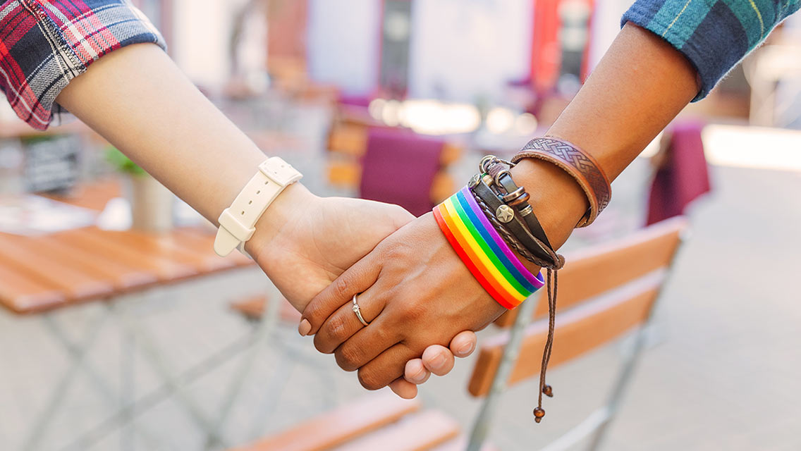 Two people, including one with a rainbow bracelet, hold hands.