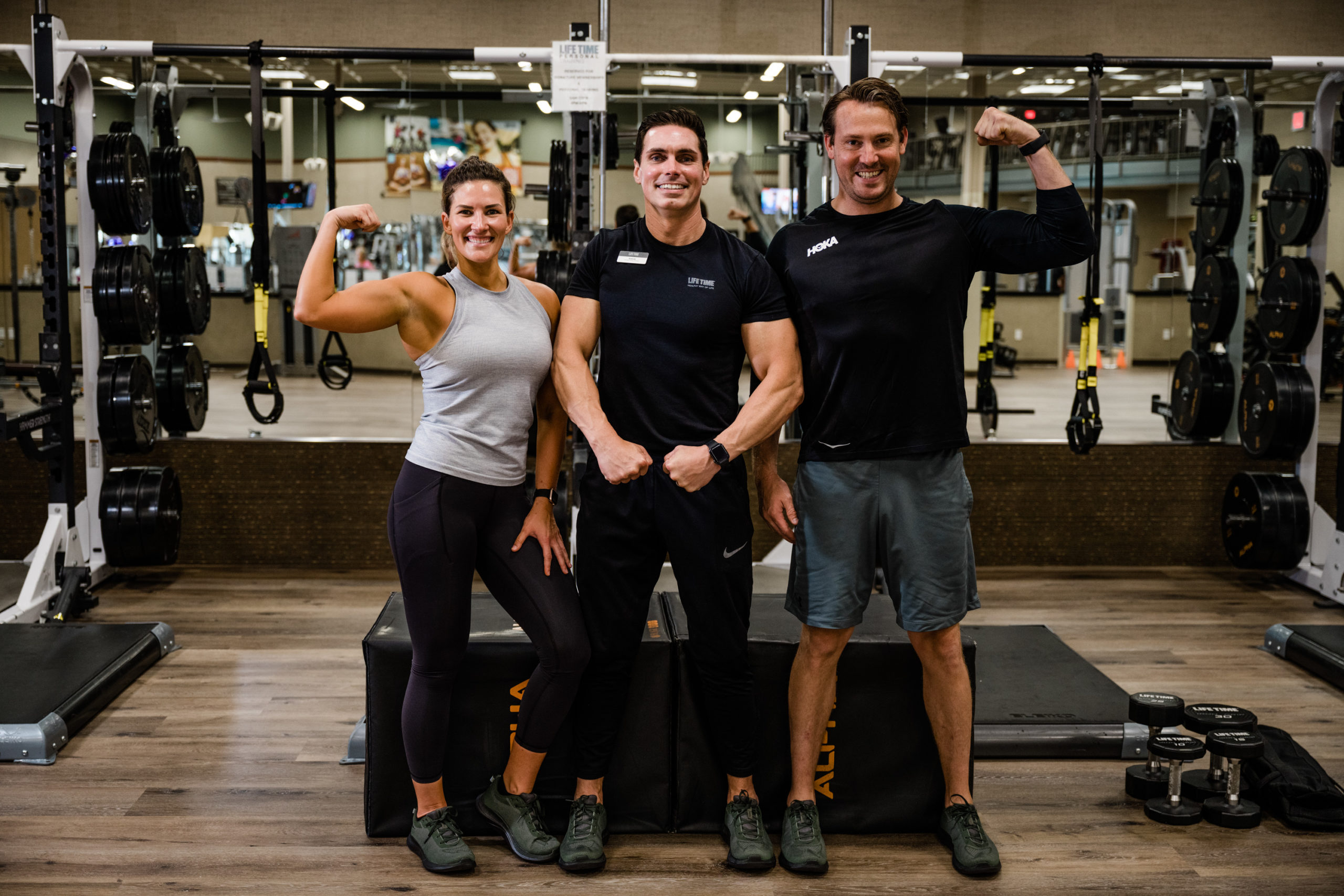 One woman and two men — all flexing — inside of a Life Time health club.