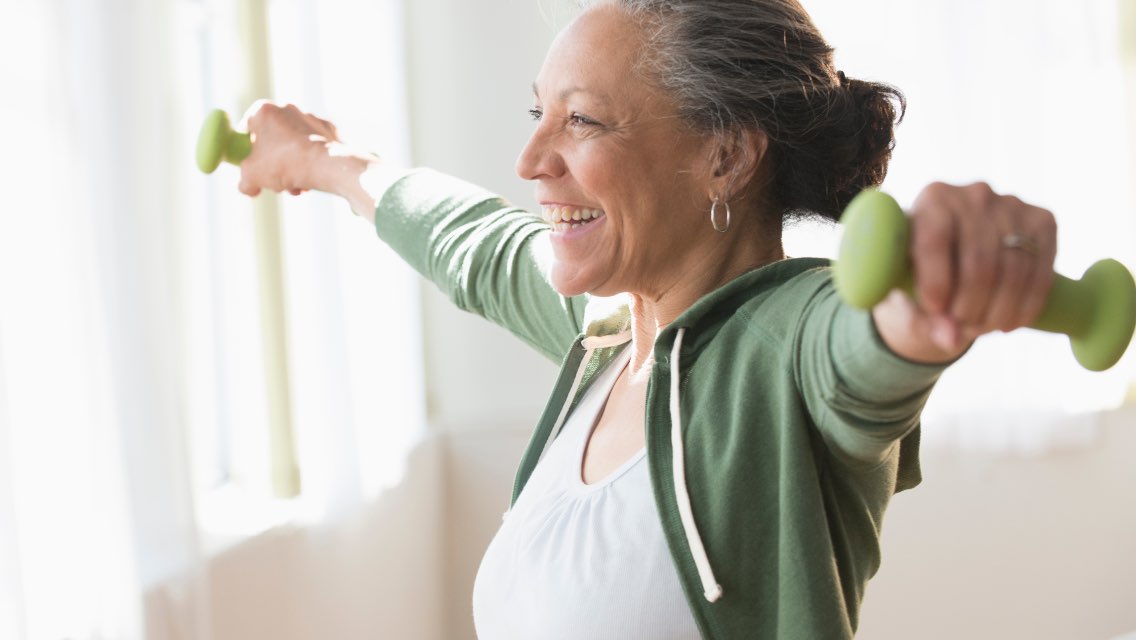An older adult woman lifting dumbbells and smiling.